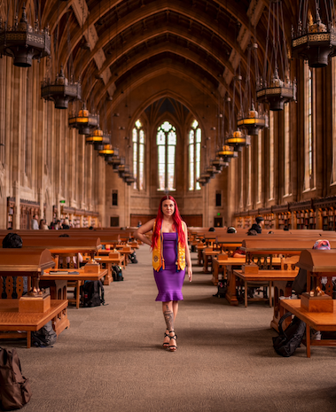 Student Bri Fero wearing a purple dress and graduation stoles and cords, in a baroque library.