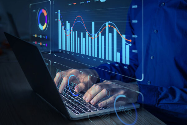 Hands typing on a keyboard. Data graphs appear on the screen.