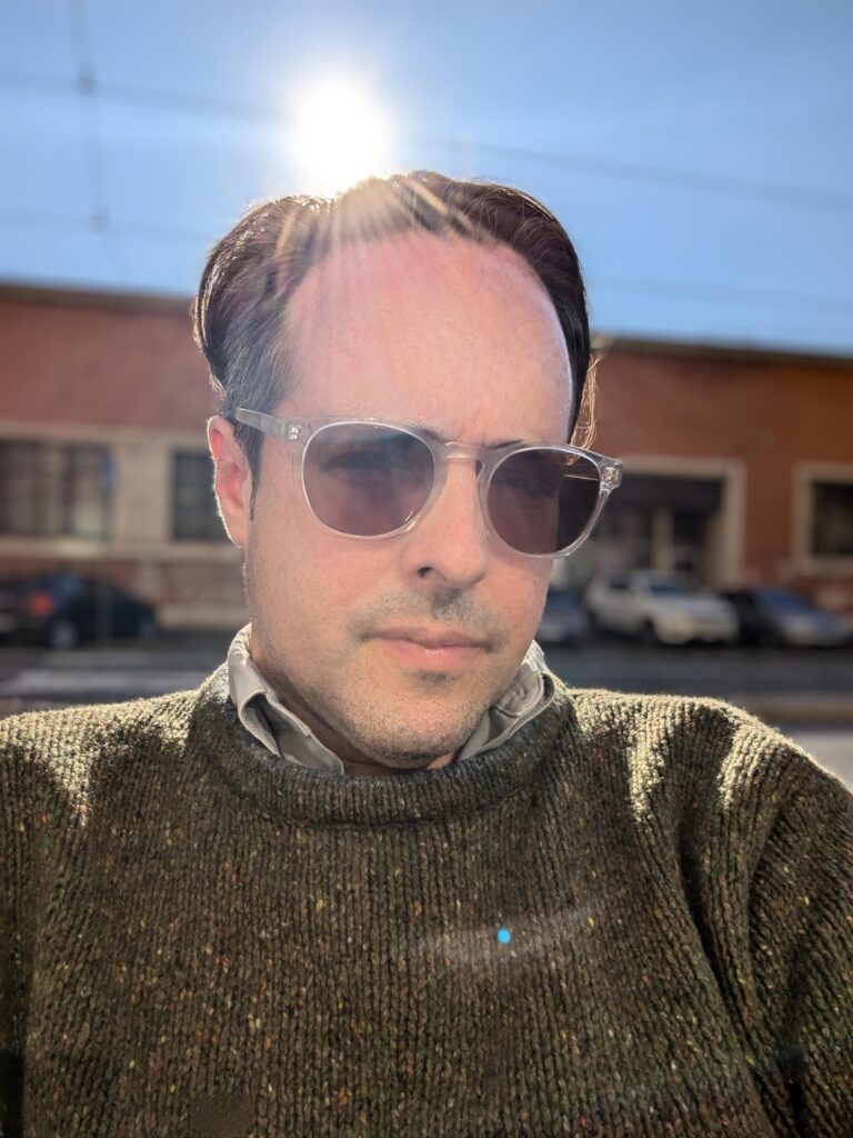 Joe Milutis is wearing sunglasses and a brown knit sweater. He is posted in front of a brick building. The sun is peaking behind in the background.