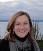 Robin Angotti with a gray scraf atop a black shirt, smiling with the water a cloudy skies in the distance